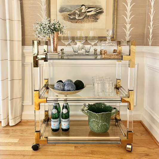 An acrylic lucite bar cart placed in a dining room corner featuring gold hardware, 3 shelves, and wheels for easy movement. The cart is decorated with flowers, spirts, glasses, and various bar accessories. 