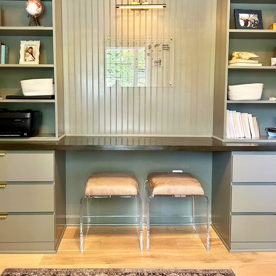 2 modern ottoman chairs with clear acrylic legs and light brown leather seat upholstery, placed underneath a counter/study area in a built-in wall bookshelf.