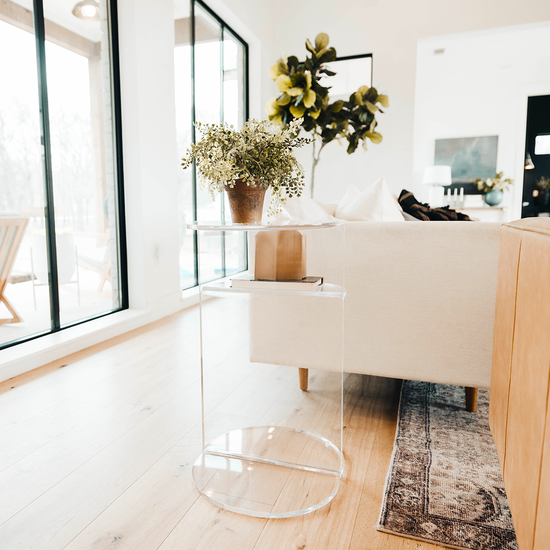 Clear acrylic side table featuring an extra shelf displaying a plant and decor beside a couch in a living room.