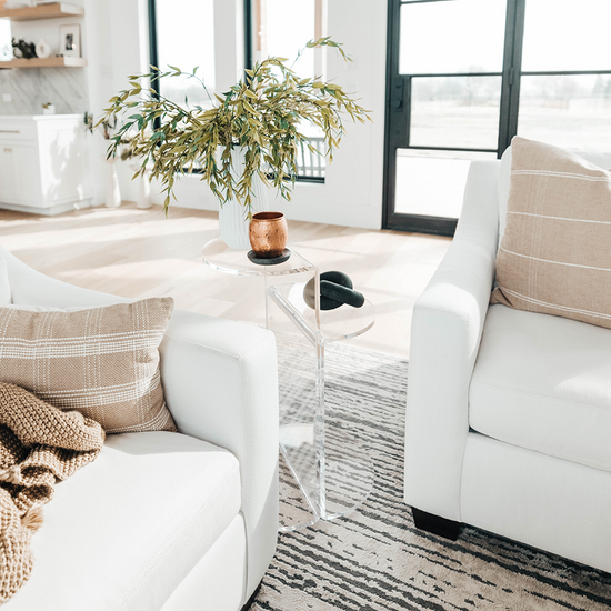 Clear acrylic side table featuring a top shelf extending 1 direction and the second shelf extending the opposite direction displaying a plant and various decor beside a couch in a living room.