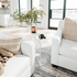 Clear acrylic side table featuring a top shelf extending 1 direction and the second shelf extending the opposite direction displaying a plant and various decor beside a couch in a living room.