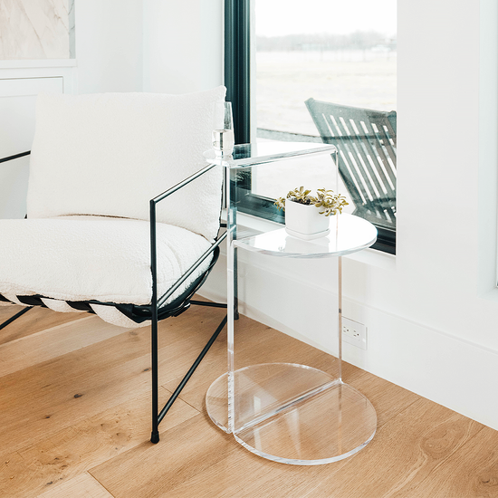 Clear acrylic side table featuring a top shelf extending 1 direction and the second shelf extending the opposite direction displaying a glass of water and a plant beside a chair next to a window.