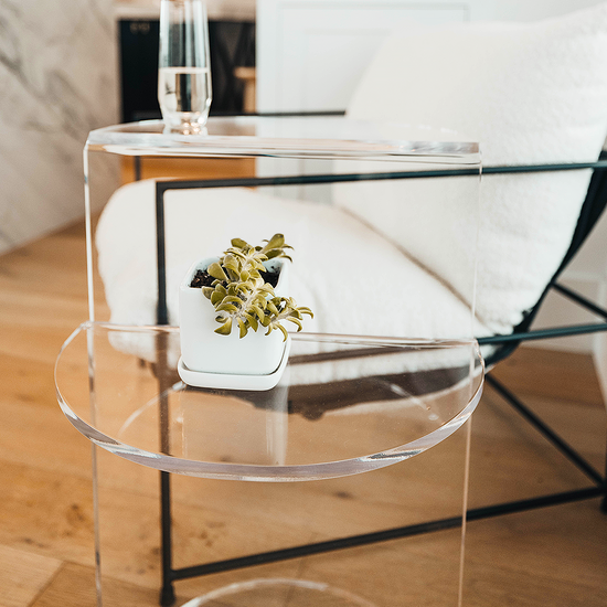 Close up of a clear acrylic side table featuring a top shelf extending 1 direction and the second shelf extending the opposite direction displaying a glass of water and a plant beside a chair.