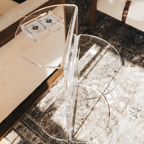 Close up of a clear acrylic side table featuring a top shelf extending 1 direction and the second shelf extending the opposite direction displaying a deck of cards in a clear acrylic card holder beside a chair.