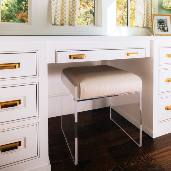 A clear acrylic accent chair with a beige upholstered seat slid underneath a white vanity with gold handles in a room with large windows and curtains.