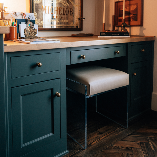 A clear acrylic accent chair with a beige upholstered seat slid underneath a dark green desk vanity with gold handles.