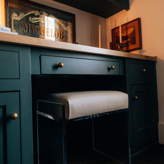 A clear acrylic accent chair with a beige upholstered seat slid underneath a dark green desk vanity with gold handles.