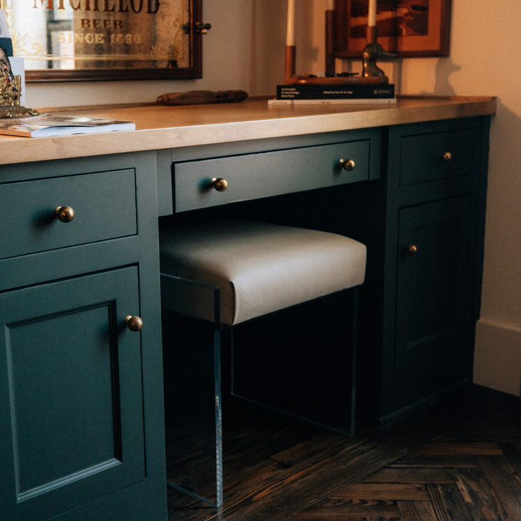 A clear acrylic accent chair with a beige upholstered seat slid underneath a dark green desk vanity with gold handles.
