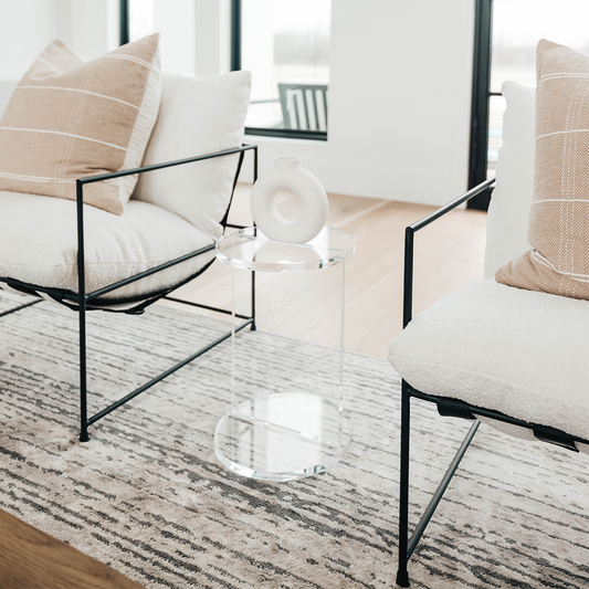 A clear acrylic lucite side table featuring a round top and round base displaying decor placed between 2 chairs in a living room. 