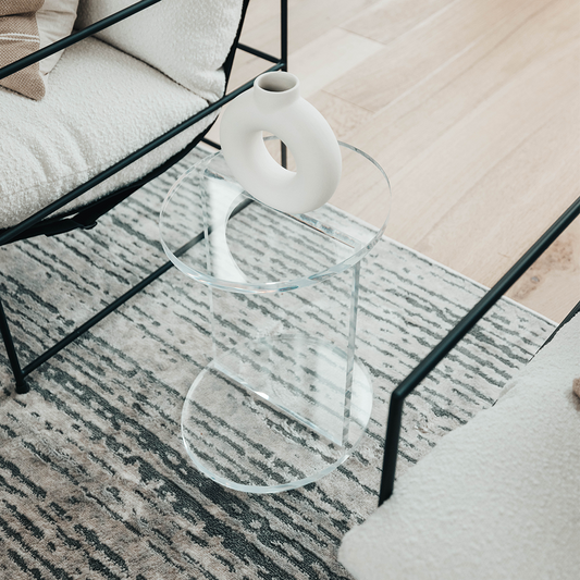 A clear acrylic lucite side table featuring a round top and round base displaying decor placed between 2 chairs in a living room. 