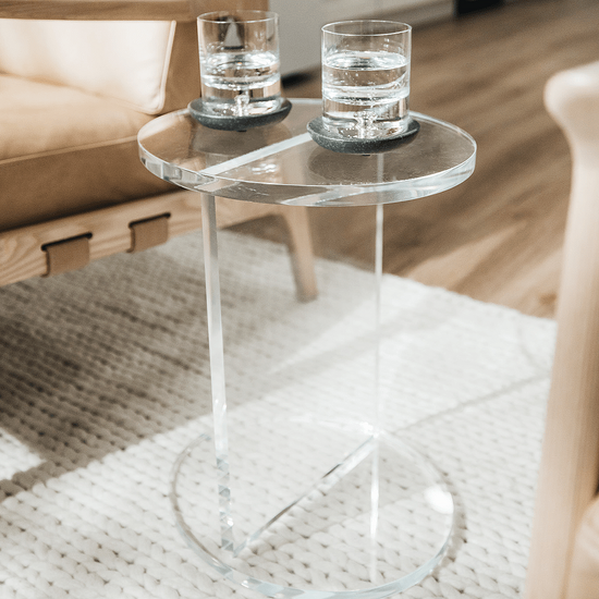 A clear acrylic lucite side table featuring a round top and round base displaying 2 glasses placed between 2 chairs in a living room. 
