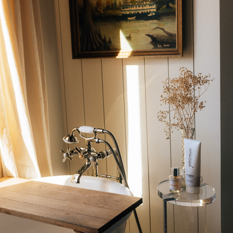 A clear acrylic lucite side table featuring a round top and round base displaying decor placed next to a soaking tub in a bathroom.