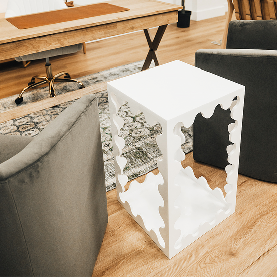 A white acrylic lucite end table featuring wavy scalloped edges placed between 2 easy chairs in an office setting.