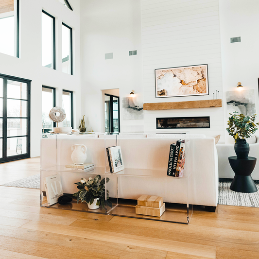 A clear acrylic lucite horizontal, 2 interior shelf bookcase displaying artwork, books, candles, and decor placed behind a sofa.