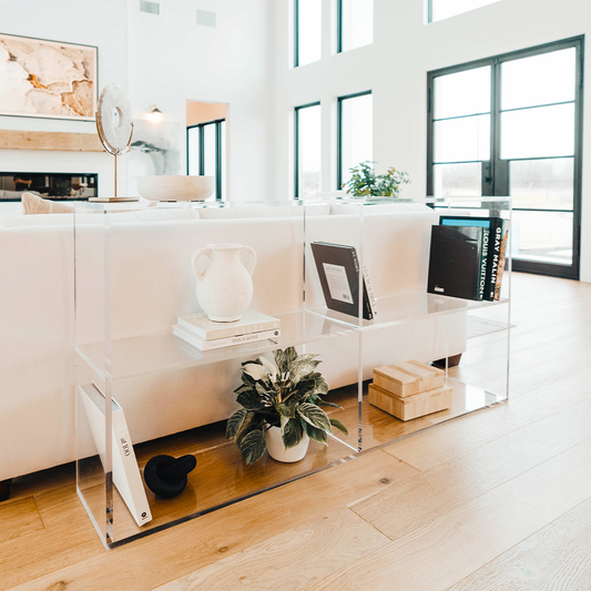 A clear acrylic lucite horizontal, 2 interior shelf bookcase displaying artwork, books, candles, and decor placed behind a sofa.