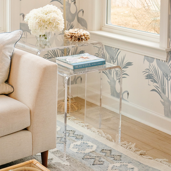 A clear acrylic nesting end table set placed in a living room setting beside a white sofa and windows.