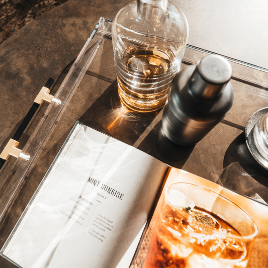 A clear acrylic lucite serving tray with gold and black handles holding a glass liquor carafe, glasses and drink receipe books placed on top of a coffee table.