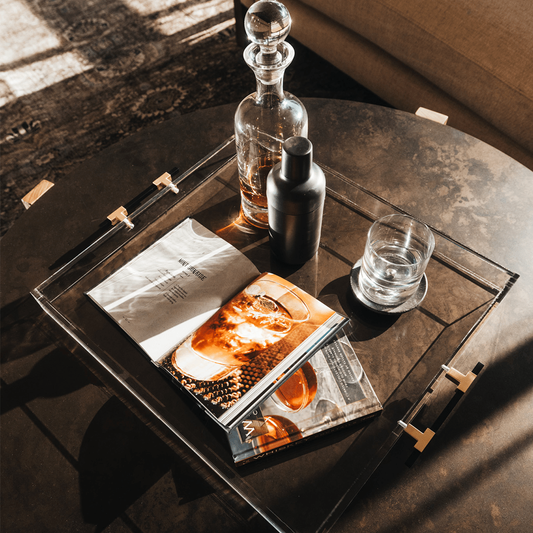 A clear acrylic lucite serving tray with gold and black handles holding a glass liquor carafe, glasses and drink receipe books placed on top of a coffee table.
