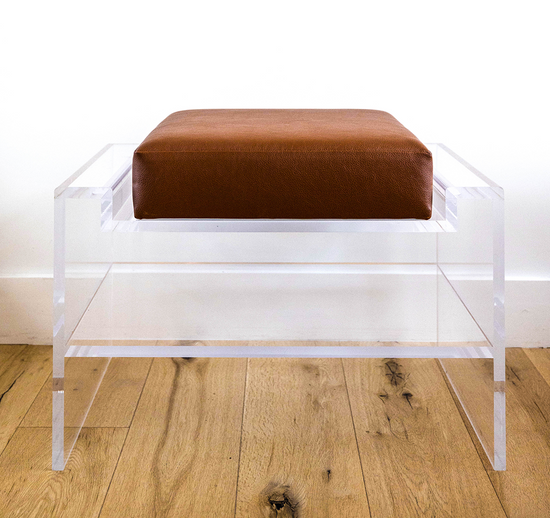 Catalog sample of a clear acrylic bench with a dark brown cushion and storage shelf underneath on a wooden floor.