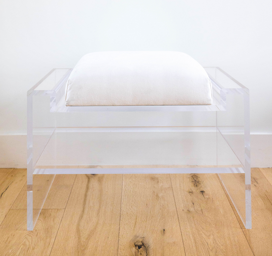 Catalog sample of a clear acrylic bench with a white cushion and storage shelf underneath on a wooden floor.