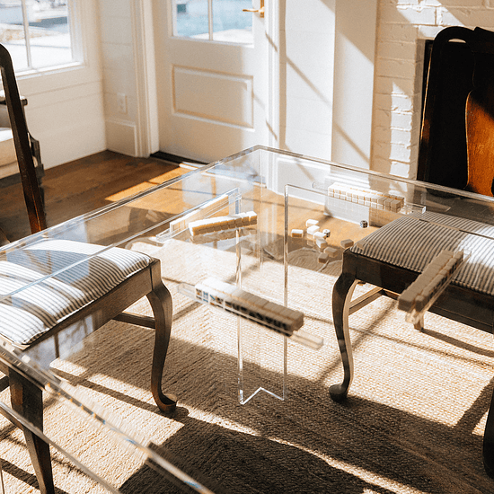 A closeup of a square clear acrylic lucite game table set up for tile game playing with seating for 1 on each side in a brightly lit living room next to a fireplace.