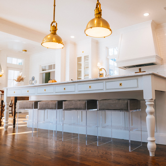 Four clear acrylic bar stools with beige velvet cushions at an white wooden island in a kitchen. 
