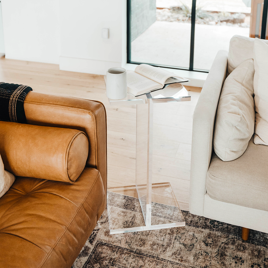 A clear acrylic side table shaped as the letter "I", positioned on a floor between a couch and an armchair displaying a coffee cup and a book. 