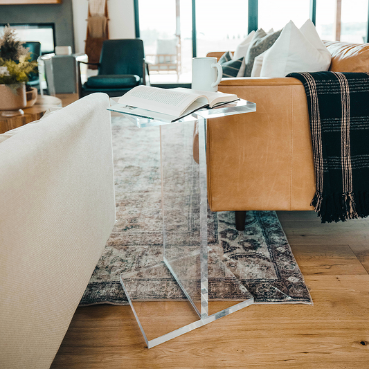 A clear acrylic side table shaped as the letter "I", known as the Dakota, positioned on a floor with a patterned rug, next to a beige sofa with a black and white checkered blanket on it.