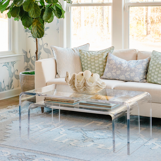 A clear acrylic lucite nesting coffee table set placed in a living room setting in front of a white sofa displaying decorative items on top and books on the smaller nesting table underneath. 
