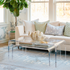 A clear acrylic lucite nesting coffee table set placed in a living room setting in front of a white sofa displaying decorative items on top and books on the smaller nesting table underneath. 