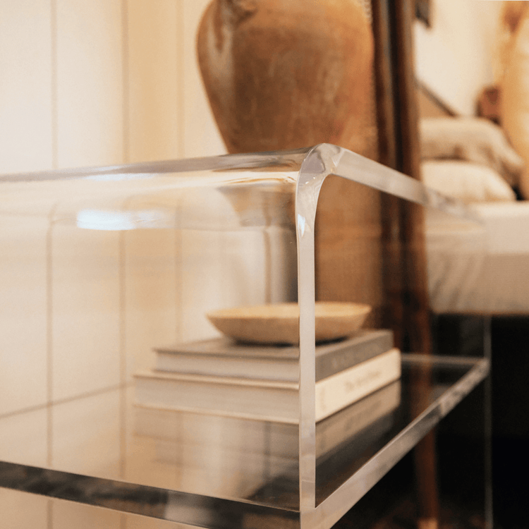 A closeup of the edges of a clear acrylic side table with an interior shelf with decorative items placed next to a bed.