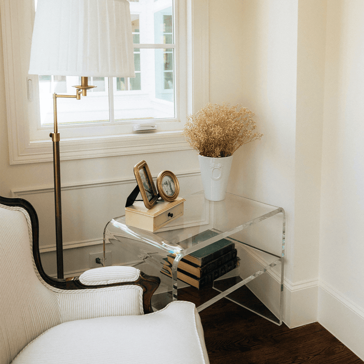 Clear acrylic side table with books and decorative items next to a white armchair in a room with a lamp and window.