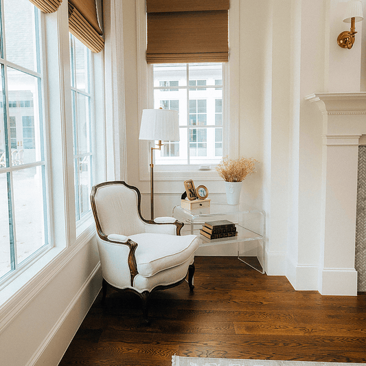 A white armchair next to a clear acrylic end table with 1 interior shelf placed in what appears to be a little reading nook in the corner of a living room with large windows.