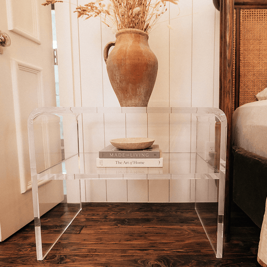 A clear acrylic side table with an interior shelf with decorative items placed next to a bed.