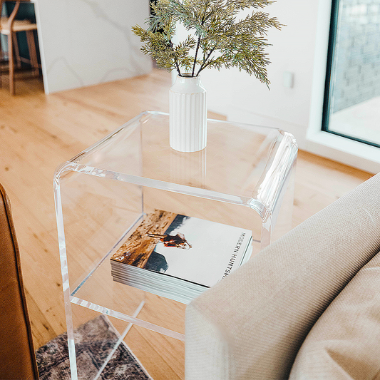 A clear acrylic lucite side table with waterfall edges and an interior shelf holding a vase of greenery and books placed between 2 couches in a living room setting. 