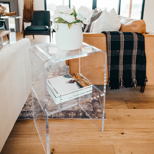 A clear acrylic lucite side table with waterfall edges and an interior shelf holding a vase of flowers and books placed between 2 couches in a living room setting. 