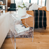A clear acrylic lucite side table with waterfall edges and an interior shelf holding a vase of flowers and books placed between 2 couches in a living room setting. 