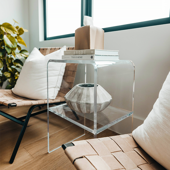 A clear acrylic lucite side table with waterfall edges and an interior shelf holding a vase and books placed beside a chair.  