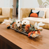 A decorative fall arrangement in a clear acrylic horizontal vase on a coffee table in a living room with beige couches and abstract wall art.