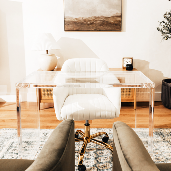 An acrylic lucite slab desk with a white chair in front, placed in an office setting with a painting on the wall and a tree in the background.
