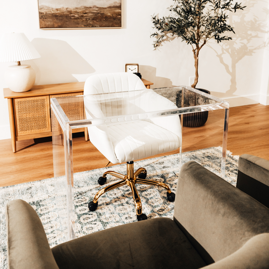 An acrylic lucite slab desk with a white chair in front, placed in an office setting with a painting on the wall and a tree in the background.