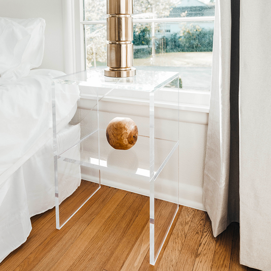Clear acrylic lucite nightstand containing 1 interior shelf with a round vase and a gold lamp on the top shelf next to a bed in a bedroom.