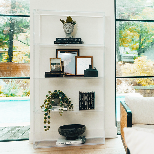 Vertical clear acrylic 4 interior shelf bookcase decorated with plants, vases, books, and decor placed between 2 large picture windows in a living room.