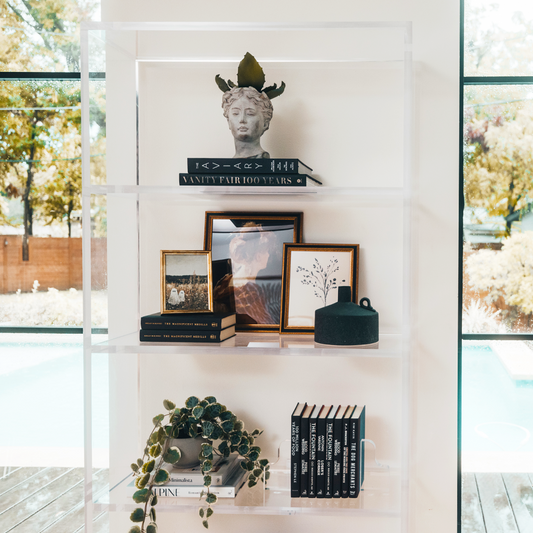 Closeup view of a vertical clear acrylic 4 interior shelf bookcase decorated with plants, vases, books, and decor placed between 2 large picture windows in a living room.