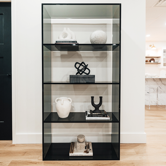 Vertical smoke gray acrylic 4 interior shelf bookcase decorated with vases, books, and decor placed against a white wall in a living room.