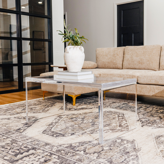 A modern acrylic lucite coffee table placed on a patterned rug, with a white vase on top of it, set against a living room backdrop with a beige couch and black door.