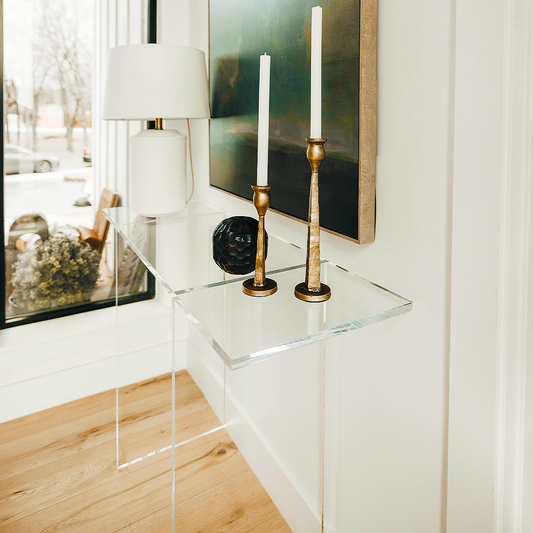 A clear acrylic lucite entryway table with a minimalist design, placed against a wall with a picture and flanked by two candlesticks on top.