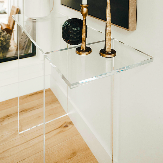 A closeup of the edges of a clear acrylic lucite entryway table with a minimalist design, placed against a wall with a picture and flanked by two candlesticks on top.