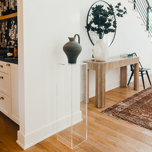 A clear acrylic lucite tall pedestal in a vertical rectangle shape placed on a wooden floor, supporting a decorative vase with flowers in an entryway setting.