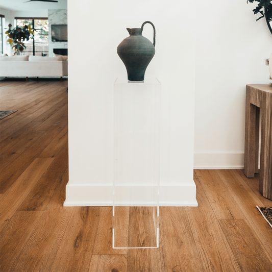 A clear acrylic lucite tall pedestal in a vertical rectangle shape placed on a wooden floor, supporting a decorative vase with flowers in an entryway setting.